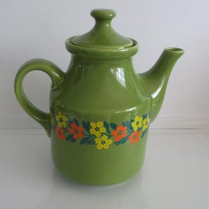 May include: A glossy olive green ceramic teapot with a matching lid, a curved handle, and a spout. The body of the teapot features a horizontal band of printed floral patterns in bright yellow and orange with dark green leaves encircling the center.