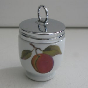 May include: White ceramic egg cup with a silver-colored lid and handle. The cup features a painted design of a red apple with green leaves. A decorative kitchenware item.