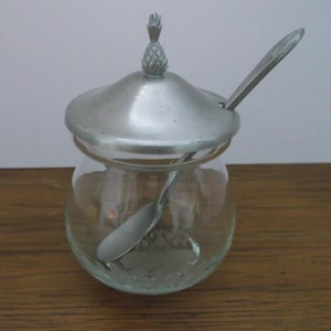 May include: A clear glass sugar bowl with a silver-colored lid and spoon. The lid is topped with a pineapple-shaped finial. The spoon rests on the rim of the bowl. The bowl has a decorative etched design.