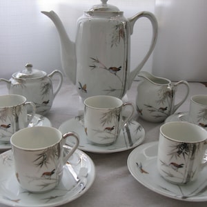 May include: A white porcelain tea set with a delicate floral design featuring birds and bamboo. The set includes a teapot, creamer, sugar bowl, and four teacups and saucers.