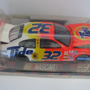 Może przedstawiać: Czerwono-biało-ż&oacute;łty model samochodu wyścigowego NASCAR z numerem 32 i logo Tide. Samoch&oacute;d znajduje się w przezroczystym plastikowym pudełku z czarną i czerwoną podstawą, na kt&oacute;rej widnieje napis "Edycja 2002".