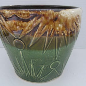 May include: A ceramic planter with a textured surface. The top section is brown and tan, while the lower half is green. The planter features a design of stylized plants and flowers.