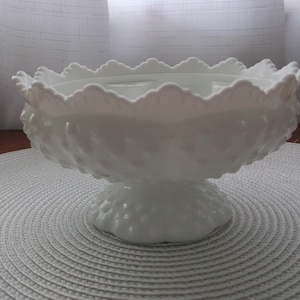 May include: White milk glass pedestal bowl with a scalloped edge and a textured design. The bowl is on a white circular placemat.