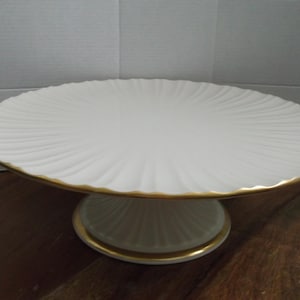 May include: A white ceramic cake stand with a gold trim. The stand has a round, ridged top and a pedestal base. The rim and base are accented with gold. The stand is designed to display cakes, pastries, or other desserts.