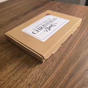 May include: A rectangular brown cardboard box featuring a white rectangular label on the top surface. The label displays the text A LITTLE BOX OF CHRISTMAS in black lettering, accompanied by a small illustration of holly leaves and a Santa hat.