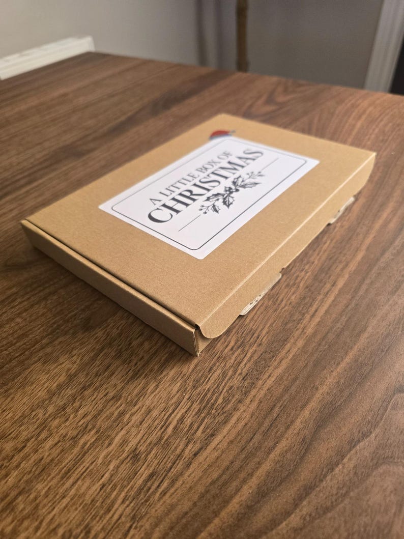 May include: A rectangular brown cardboard box featuring a white rectangular label on the top surface. The label displays the text A LITTLE BOX OF CHRISTMAS in black lettering, accompanied by a small illustration of holly leaves and a Santa hat.