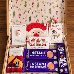 May include: A cardboard gift box lined with Christmas-themed paper containing two Cadbury instant hot chocolate sachets, a Walkers shortbread fingers 40g pack, a Cadbury Dairy Milk Little Bar, a crochet Santa face, and three holiday cards.