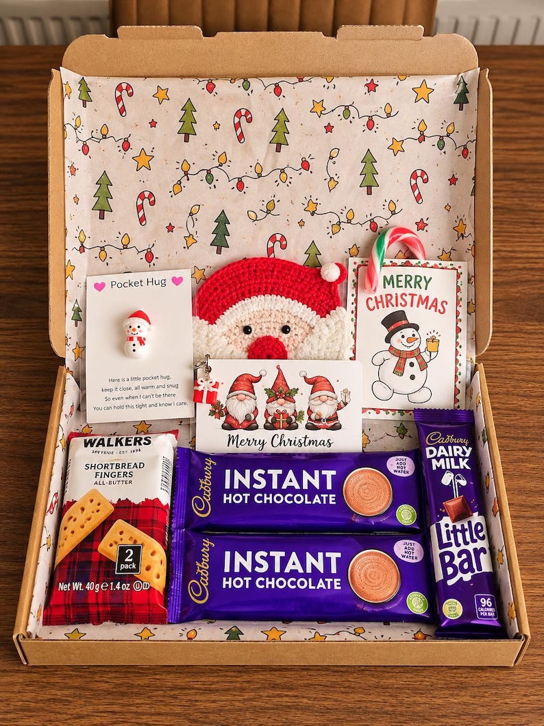 May include: A cardboard gift box lined with Christmas-themed paper containing two Cadbury instant hot chocolate sachets, a Walkers shortbread fingers 40g pack, a Cadbury Dairy Milk Little Bar, a crochet Santa face, and three holiday cards.