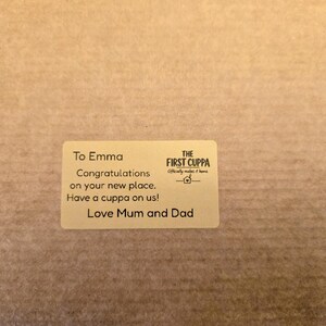 May include: A rectangular beige sticker featuring black text which reads: To Emma, Congratulations on your new place. Have a cuppa on us! Love Mum and Dad. The logo for The First Cuppa, including a small house icon, is printed on the right-hand side.