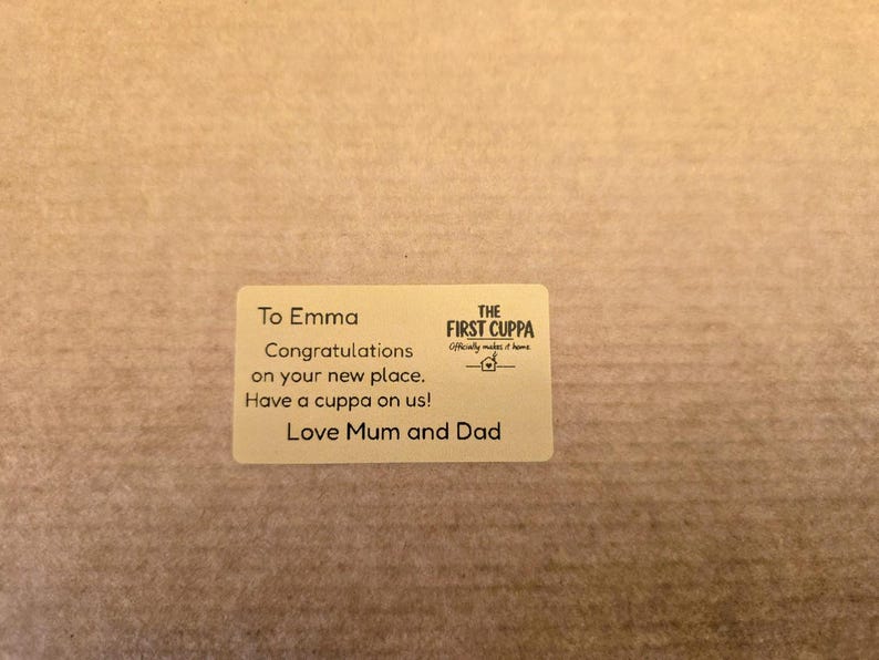 May include: A rectangular beige sticker featuring black text which reads: To Emma, Congratulations on your new place. Have a cuppa on us! Love Mum and Dad. The logo for The First Cuppa, including a small house icon, is printed on the right-hand side.