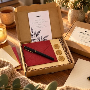 May include: A stationery kit in a cardboard box containing a black fountain pen, a deep red envelope, three gold wax seals with leaf imprints, and a card with the text NOT SURE WHERE TO START? A FEW IDEAS FOR PUTTING PEN TO PAPER.