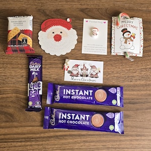May include: A holiday gift set containing a Walkers shortbread fingers pack, a crocheted Santa face, a Pocket Hug token, a candy cane with a snowman card, a gnome gift tag, a Cadbury Dairy Milk bar, and two Cadbury instant hot chocolate sachets.