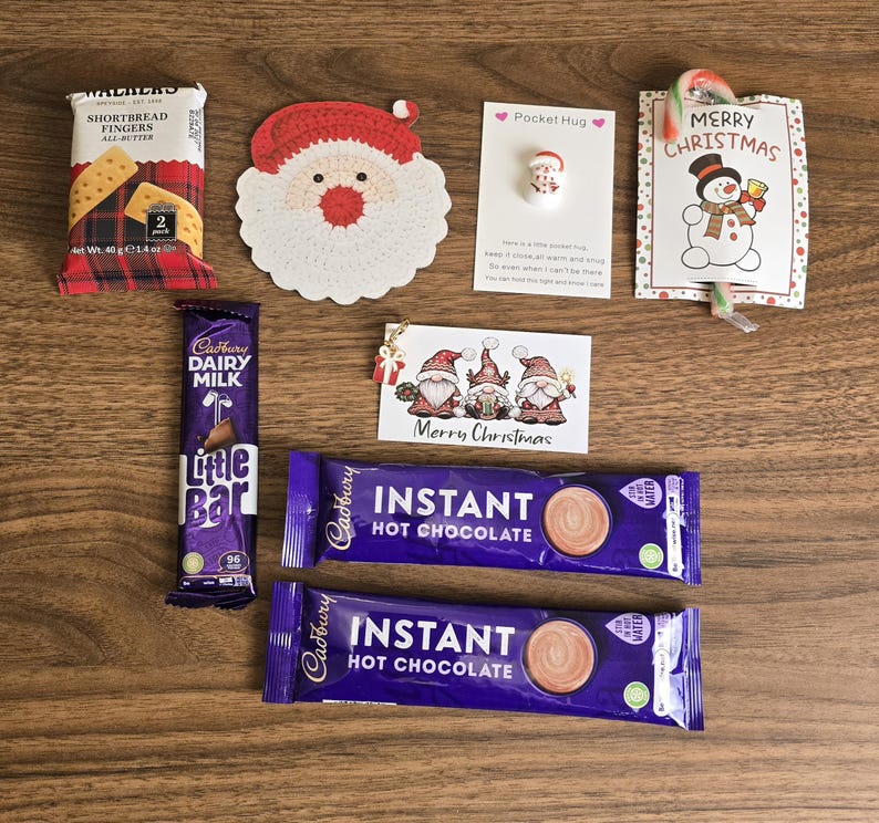 May include: A holiday gift set containing a Walkers shortbread fingers pack, a crocheted Santa face, a Pocket Hug token, a candy cane with a snowman card, a gnome gift tag, a Cadbury Dairy Milk bar, and two Cadbury instant hot chocolate sachets.