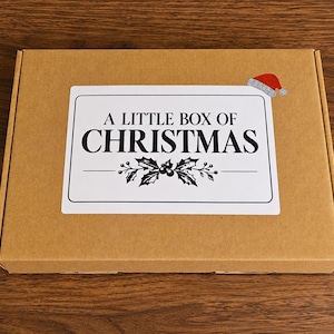 May include: A rectangular brown cardboard box featuring a white rectangular label on the top surface. The label displays the black text A LITTLE BOX OF CHRISTMAS, a graphic of holly leaves and berries, and a small red Santa hat icon.