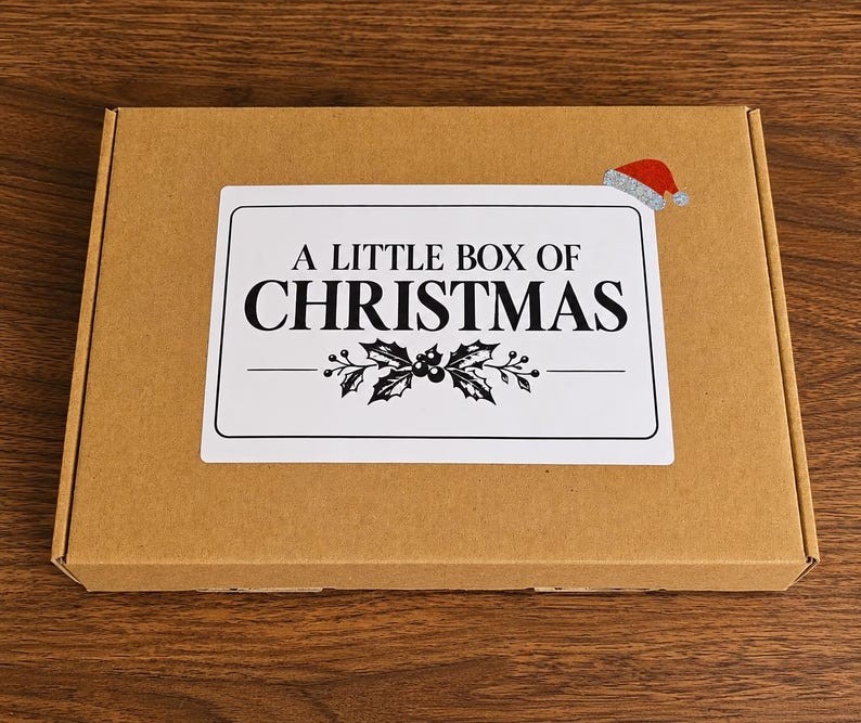 May include: A rectangular brown cardboard box featuring a white rectangular label on the top surface. The label displays the black text A LITTLE BOX OF CHRISTMAS, a graphic of holly leaves and berries, and a small red Santa hat icon.