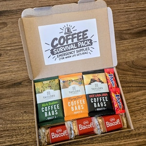 May include: A cardboard gift box containing three Taylors of Harrogate coffee bag packets in green, orange, and black, two red Daim chocolate bars, and three Lotus Biscoff biscuit packs. The lid features a label reading Coffee Survival Pack.