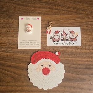 May include: A crochet Santa Claus face coaster, a Pocket Hug card with a small snowman charm, and a Merry Christmas card featuring three gnomes and a small gift charm with a gold lobster clasp, all arranged on a wooden surface.
