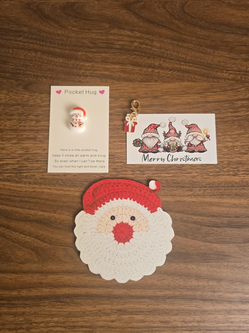 May include: A crochet Santa Claus face coaster, a Pocket Hug card with a small snowman charm, and a Merry Christmas card featuring three gnomes and a small gift charm with a gold lobster clasp, all arranged on a wooden surface.