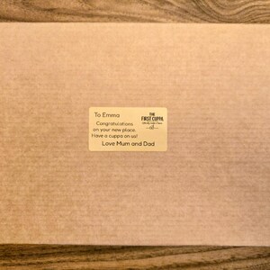 May include: A rectangular brown cardboard shipping box featuring a centered rectangular sticker. The sticker displays the text: To Emma, Congratulations on your new place. Have a cuppa on us! Love Mum and Dad, and the logo for The First Cuppa.