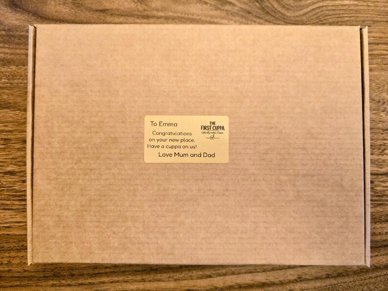 May include: A rectangular brown cardboard shipping box featuring a centered rectangular sticker. The sticker displays the text: To Emma, Congratulations on your new place. Have a cuppa on us! Love Mum and Dad, and the logo for The First Cuppa.