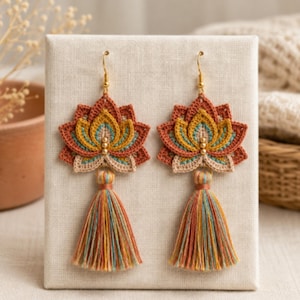 May include: A pair of dangle earrings featuring a crocheted lotus flower design in shades of rust, mustard yellow, teal, and cream. Each earring is accented with three small gold beads and finished with a multi-colored thread tassel at the base.