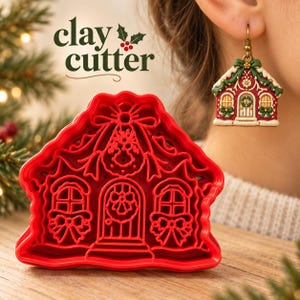 Puede incluir: Un cortador de arcilla de plástico rojo con forma de casa con detalles intrincados que incluyen una puerta central, ventanas y lazos. El texto clay cutter es visible arriba. Se muestra un pendiente a juego con forma de casa.
