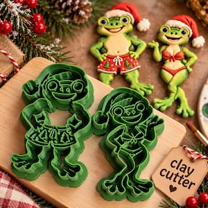May include: Two green plastic cookie cutters shaped like frogs wearing Santa hats. One frog wears patterned shorts, the other a bikini. Beside them are two matching frog cookies. A brown tag with the text clay cutter is attached.