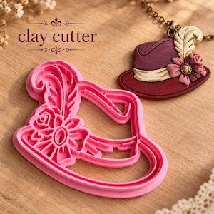 Könnte beinhalten: Ein rosafarbener Kunststoff-Ausstecher in Hutform mit Feder- und Blumendetail, begleitet von einem fertigen dunkelvioletten Hut-Anhänger an einer Goldkette. Der Text "clay cutter" ist dunkelbraun über dem Ausstecher gedruckt.
