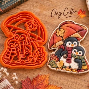 May include: An orange plastic clay cutter and a corresponding finished clay figure featuring two penguins standing under an umbrella, surrounded by autumn leaves. The penguins wear scarves, and the text Clay Cutter is visible above them.