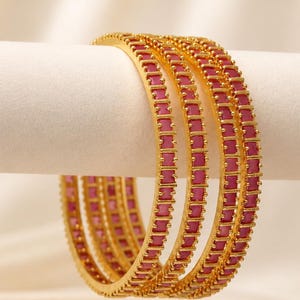May include: A set of three gold-toned bangles, each featuring a continuous row of rectangular, deep pink stones set in a metallic frame. The bangles have a polished finish and a uniform design, showcasing a series of small, prong-set stones.