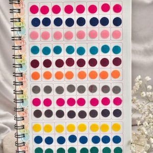May include: A spiral-bound book titled Ananya Kumkum Bindi featuring twelve horizontal rows of eight circular adhesive bindis in various solid colors including pink, navy, light pink, teal, purple, orange, gray, magenta, black, yellow, blue, and green.