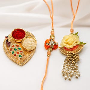May include: A gold-toned heart-shaped tray featuring a peacock design with red and green accents, holding two small bowls of red powder and white rice, alongside two decorative wrist bands with floral, bead, and crystal embellishments.