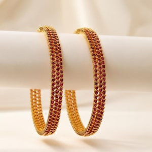 May include: Two circular gold bangles featuring a textured, lattice-like metal frame embedded with numerous small, deep red stones. The bangles have a consistent, repeating pattern of gold metal and red stones arranged in a grid-like structure.