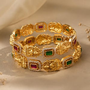 Puede incluir: Dos brazaletes dorados con motivos de cabezas de león en relieve que alternan con engastes rectangulares. Cada engaste contiene una piedra preciosa verde esmeralda o roja rubí rodeada por pequeñas piedras blancas brillantes.