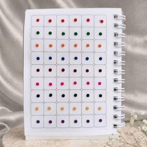 May include: A white spiral-bound notebook featuring a grid of rounded squares on the cover. Each square contains a small, solid-colored dot. The dots are arranged in horizontal rows, each row displaying a different color, including red, green, orange, blue, maroon, pink, black, yellow, and purple.