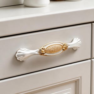 May include: A horizontal drawer pull featuring a central oval medallion with a textured gold finish, surrounded by a white frame. The handle arms extend outward with a scalloped, fan-like design in a matte white finish, attached to a drawer.