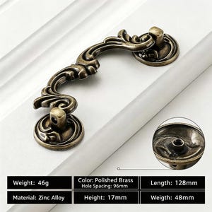May include: A polished brass drawer pull with an ornate, curved design and two circular mounting bases. The hardware has a length of 128mm, a height of 17mm, and a hole spacing of 96mm. The zinc alloy handle weighs 46g to 48g.
