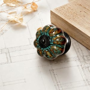 Puede incluir: Un tirador de mueble con forma de calabaza que presenta un esmalte brillante y jaspeado en tonos marrón oscuro y turquesa. El centro del tirador tiene una placa metálica circular negra con un borde estriado y un pequeño tornillo central.