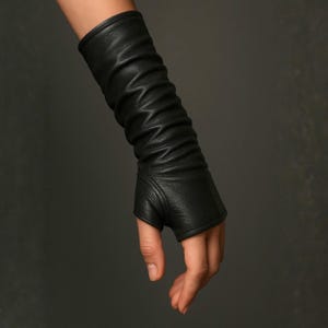 May include: A black leather fingerless glove extending to the forearm, featuring a textured surface with gathered, ruched detailing along the length and a reinforced seam around the thumb opening, providing a snug fit from the wrist to the mid-forearm.