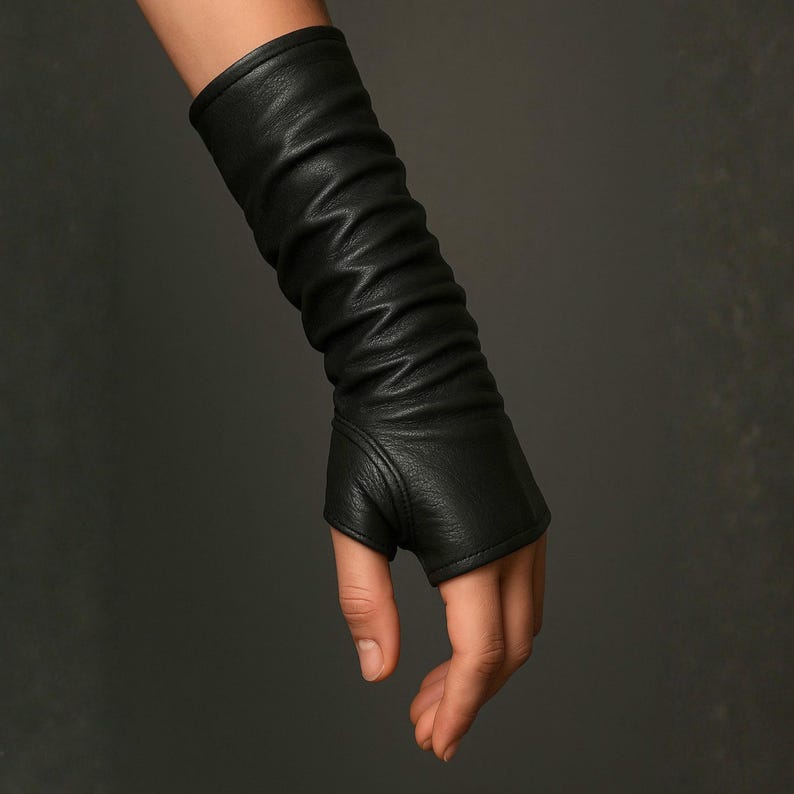 May include: A black leather fingerless glove extending to the forearm, featuring a textured surface with gathered, ruched detailing along the length and a reinforced seam around the thumb opening, providing a snug fit from the wrist to the mid-forearm.