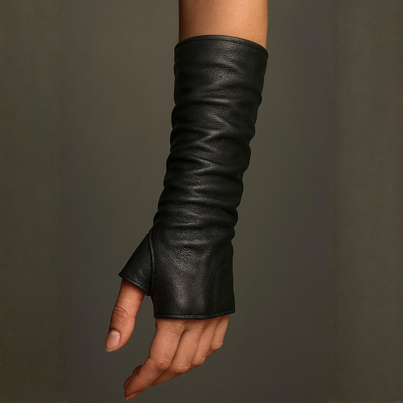 May include: A long black leather glove that extends past the wrist and up the forearm. The glove features a thumb hole and a fingerless design, with a soft, textured surface that creates natural folds and creases along the length of the arm.