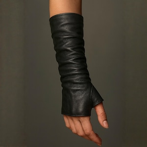 May include: A black leather fingerless glove extending to the forearm, featuring a textured, slightly ruched surface and a dedicated opening for the thumb. The design covers the wrist and lower arm, leaving the fingers exposed for full dexterity.