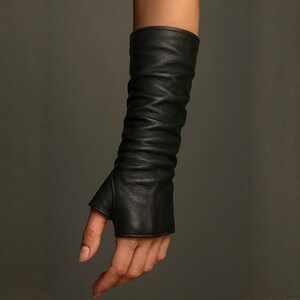 May include: A long black leather glove that extends past the wrist and up the forearm. The glove features a thumb hole and a fingerless design, with a soft, textured surface that creates natural folds and creases along the length of the arm.