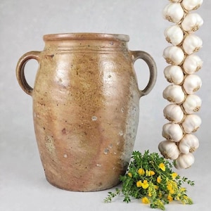 Peut inclure: Un grand pot en céramique cylindrique de couleur beige avec deux anses latérales et une surface texturée et tachetée. À côté du pot est suspendue une longue tresse verticale de bulbes d'ail blancs, accompagnée d'un bouquet d'herbes vertes à fleurs jaunes.