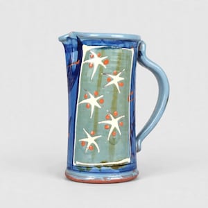 Op de afbeelding: Een keramische kan met een cilindrisch lichaam, een lichtblauw handvat en een donkerblauwe rand. Het centrale paneel is saliegroen, versierd met witte stervormige motieven en kleine oranje cirkelvormige accenten. De basis toont roodbruine klei.