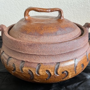 May include: A round, brown, textured ceramic pot with a matching lid and two side handles. The exterior features a horizontal band of carved, dark, crescent-shaped indentations around the lower section, with a smooth, arched handle on the lid.