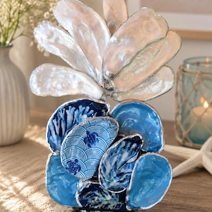Puede incluir: Una escultura decorativa compuesta por múltiples conchas de ostra dispuestas en un grupo vertical. Las conchas superiores son de color blanco iridiscente, mientras que las inferiores presentan varios tonos de azul, incluyendo patrones de tortugas marinas y coral.