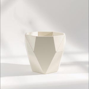 May include: A matte off-white planter featuring a faceted, geometric exterior with multiple angular planes. The vessel has a wide, open top and a tapered base, showcasing a structured, multi-sided design that creates distinct light and shadow.