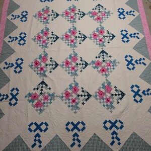 May include: A rectangular quilt featuring a cream-colored background with a repeating pattern of diamond-shaped blocks. Each block contains a grid of pink, light blue, and dark blue squares. The quilt is bordered by a pink fabric edge.