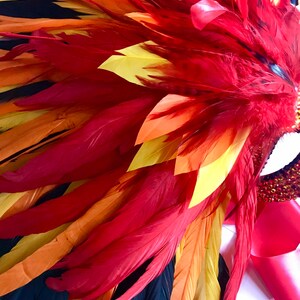 May include: A vibrant headdress featuring a cascade of red, orange, yellow, and black feathers. The feathers are arranged in a layered pattern, creating a dynamic and eye-catching design. A red ribbon and jeweled accents are also visible.
